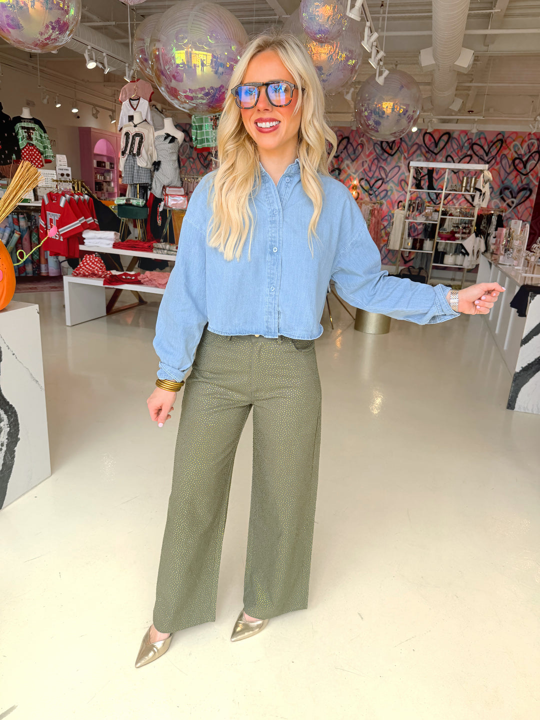 A woman posing in a boutique store wearing a light blue long-sleeve button-up shirt, olive green wide-leg pants, and metallic gold pointed heels